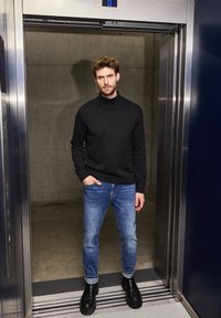 Svart polotröja, blå slim-fit jeans och svarta ankelskor. Miljön är en hiss med metalliska accenter.