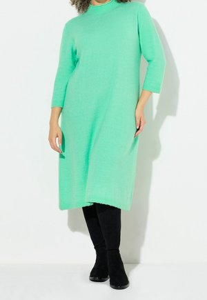 Kvinde iført en mintgrøn lang sweaterkjole med trekvart ærmer og sorte knæhøje støvler, stående mod en hvid baggrund.