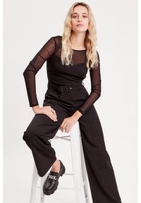 Combinaison noire avec un buste ajusté, de longues manches transparentes à pois, une taille ceinturée et des jambes larges. Associée à des mocassins noirs avec un accent en chaîne argentée.