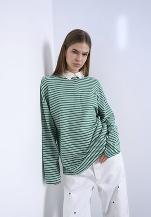 Kvinde med langt hår iført en grøn- og hvidstribet sweater med en hvid krave, lænende sig op ad en hvid væg, hænderne i lommerne på hvide bukser.