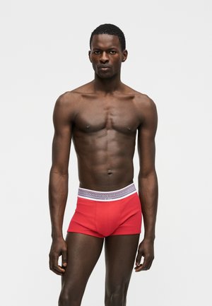 Homme athlétique debout portant un boxer rouge avec une ceinture blanche ornée d'un motif géométrique noir et du logo Versace.