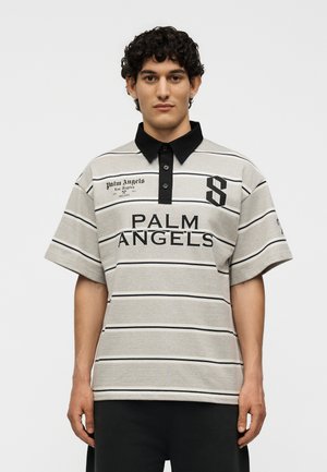 Άνδρας με σγουρά μαλλιά, φορώντας γκρι και μαύρο ριγέ πόλο πουκάμισο με το κείμενο "PALM ANGELS" και μαύρο γιακά, στέκεται μπροστά σε απλό φόντο.