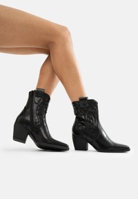 Botas de cuero negro hasta el tobillo con puntas afiladas, tacones cuadrados, patrones en relieve en la parte superior y cierre de cremallera lateral. Acabado suave en todo.