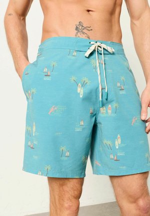 Mann trägt türkisfarbene Badeshorts mit Mustern aus Segelbooten, Palmen, Surfbrettern und Hummern, steht mit den Händen in den Taschen.