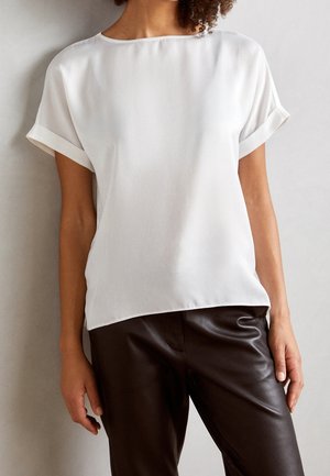 Donna che indossa una blusa bianca a maniche corte con polsini arrotolati e pantaloni in pelle marrone scuro, in piedi contro uno sfondo chiaro e neutro.