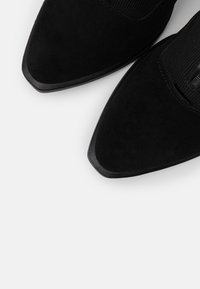 Botines de ante negros con puntas afiladas, paneles laterales elásticos y suelas de goma negra lisa. La textura parece suave y sutil.