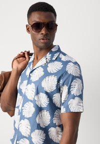 Korte mouwen blauwe shirt met een wit bladdessin, knoopsluiting aan de voorkant en een ontspannen kraag. Model houdt een bruine tas vast en draagt een zonnebril.