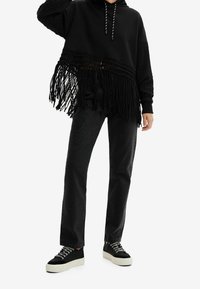 Sudadera negra con dobladillo desflecado y mangas texturizadas, combinada con jeans negros con adornos y zapatillas negras con suelas blancas.