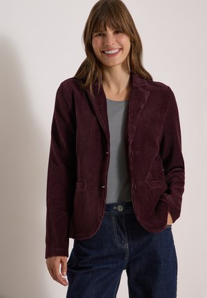 Femme souriante portant un blazer en velours côtelé violet foncé par-dessus une chemise grise et un jean bleu, debout devant un fond clair uni.
