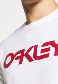 Bílý bavlněný tričko s výrazným červeným logem "OAKLEY". Kulatý výstřih a krátké rukávy. Zblízka se zdůrazňují designové detaily.