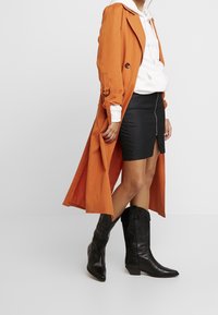 Oranžový dlouhý kabát s předními knoflíky, bílá mikina s kapucí, černá mini sukně se zipem a černé kovbojské boty. Hladká textura a vrstvený design.
