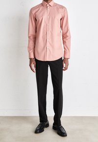 Lichtroze overhemd met puntige kraag, gecombineerd met zwarte pantalon en zwarte loafers, tegen een neutrale achtergrond.