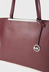 Burgundská kožená taška přes rameno s texturovaným povrchem, stříbrným kováním a kovovým logem na přívěsku. Disponuje pevnými, dlouhými uchy pro nošení.