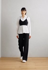 Top cropped nero sopra una camicia bianca con bottoni, abbinato a pantaloni neri larghi e sneakers bianche. Sfondo con pavimento in legno e parete grigia.