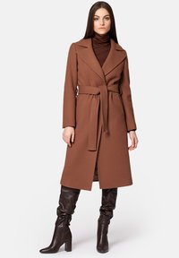 Brązowy wełniany płaszcz z paskiem w talii, klapowanym kołnierzem i długimi rękawami. Noszony na ciemnym golfie, w połączeniu z brązowymi botkami do kolan.