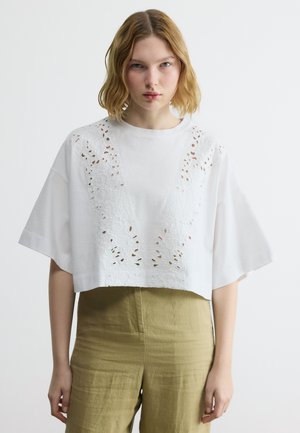 Νεαρή γυναίκα με ξανθά μαλλιά μήκους έως τους ώμους, που φοράει ένα λευκό μπλουζάκι με κοντά μανίκια και λεπτομέρειες από δαντέλα, καθώς και χακί ψηλόμεσο παντελόνι.