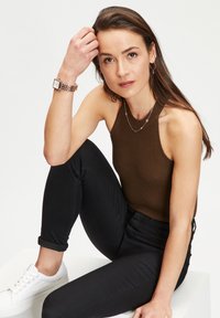 Braun gerippter Tanktop, schwarze enge Jeans, weiße Sneakers. Kombiniert mit geschichteten silbernen Halsketten und einer quadratischen Uhr mit Metallarmband.