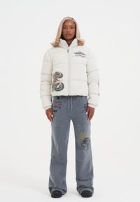 Donna in piedi che indossa una giacca a vento bianca con toppe colorate e pantaloni grigi a gamba larga con stampe grafiche.