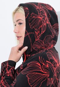 Felpa nera con cappuccio decorata da vivaci motivi floreali rossi. Presenta una chiusura con zip e un tessuto testurizzato, indossata da un modello con una mano vicino al cappuccio.
