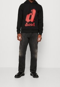 Svart huvtröja med en stor röd 'd'-logotyp och texten "diesel", kombinerad med mörkgrå jeans och svarta snörskor.