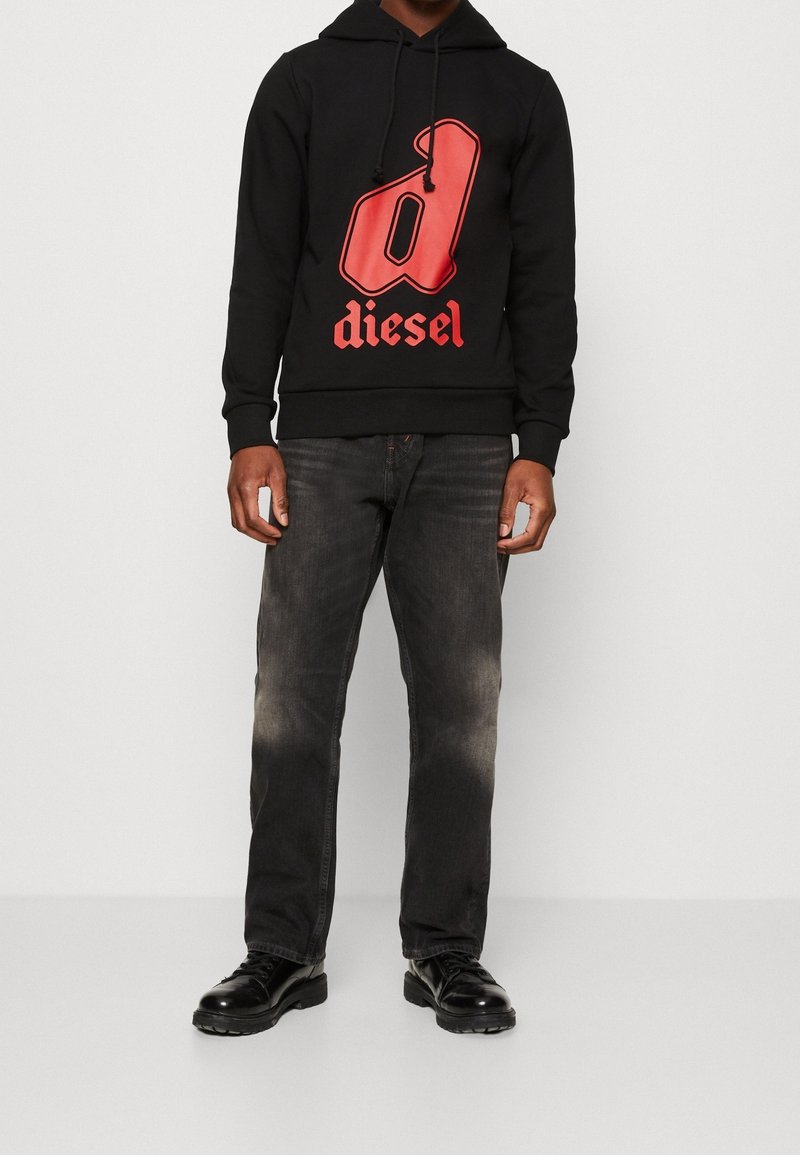 Svart huvtröja med en stor röd 'd'-logotyp och texten "diesel", kombinerad med mörkgrå jeans och svarta snörskor.