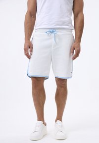 Pantalones cortos deportivos de color gris claro con acentos en azul claro, cinturilla con cordón, bolsillos laterales y dobladillo curvado. Llevados con zapatillas blancas.