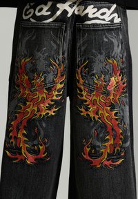 Schwarze Denim-Jeans mit lebendigen roten und gelben flammenden Phönix-Designs auf den Gesäßtaschen unter dem weißen stilisierten Textbund.