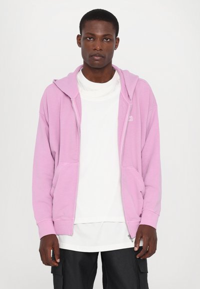 Pink zip-up hoodie with a hood, featuring front pockets and ribbed cuffs. Worn over a white shirt, paired with dark pants. Smooth texture.