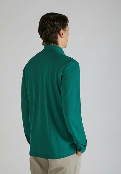 Camicia verde a maniche lunghe in tessuto liscio, con colletto e polsini abbottonati, abbinata a pantaloni beige. Vista posteriore mostrata.