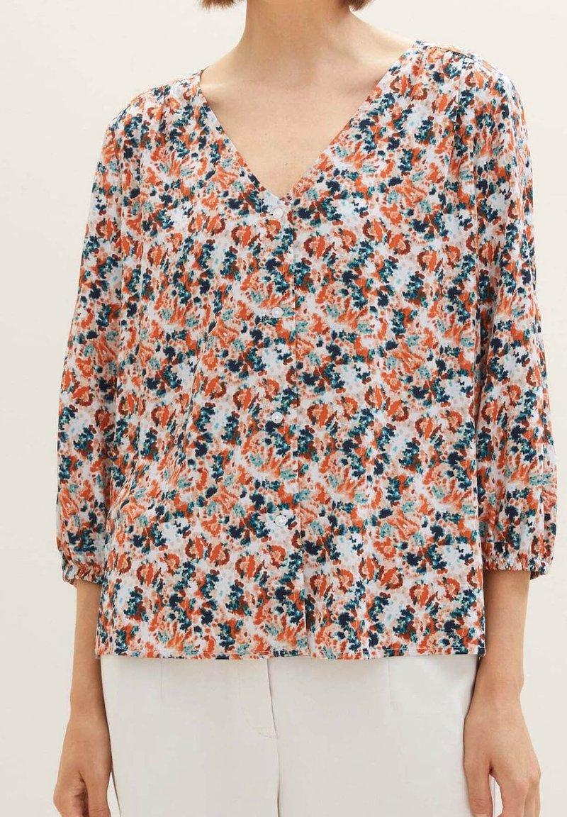 Femme portant un chemisier boutonné à manches trois-quarts, avec un imprimé floral abstrait orange, turquoise et marine sur fond blanc.