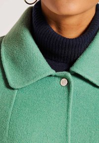 Groene wolmixjas met een platte kraag, voorzien van een zilveren knoopdetail. Onderaan draagt de model een marineblauwe geribbelde coltrui.