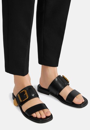 Sandalias de cuero negro con dos correas anchas, una de ellas con un acento de hebilla dorada. Suela plana y diseño abierto para mayor comodidad.
