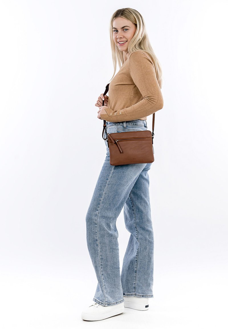 Borsa a tracolla in pelle marrone con tasca anteriore con zip, texture morbida e tracolla regolabile, indossata con jeans blu a vita alta.