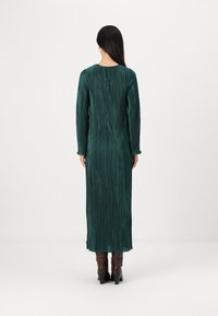 Πράσινο πλισέ φόρεμα με μακριά μανίκια και στρογγυλή λαιμόκοψη, διαθέτοντας ρέουσα σχεδίαση που φτάνει έως τους αστραγάλους, συνδυασμένο με καφέ μπότες.