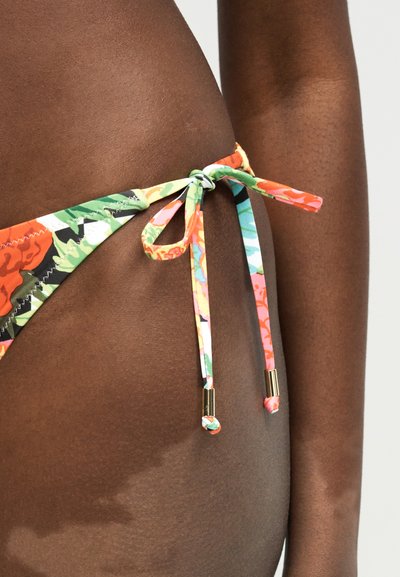 Πλάγια όψη ενός σκούρου δέρματος ατόμου που φοράει μπικίνι με λουλουδάτο κάτω μέρος δεμένο με φιόγκο με κορδόνια με χρυσά καπάκια στον γοφό.