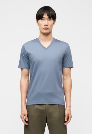 Tânăr cu păr negru drept, purtând un tricou cu mâneci scurte, de culoare albastru deschis, cu decolteu în V, și pantaloni verde măsliniu, stând în picioare pe un fundal alb.