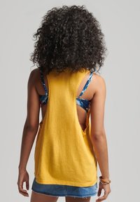 Top giallo senza maniche con ampie scollature, realizzato in tessuto morbido, indossato su un reggiseno multicolore a fantasia, abbinato a una gonna di jeans.