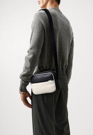 Torebka crossbody w granatowo-kremowej skórze, prostokątna, z zapięciem na zamek błyskawiczny i regulowanym paskiem, noszona do szarego swetra.
