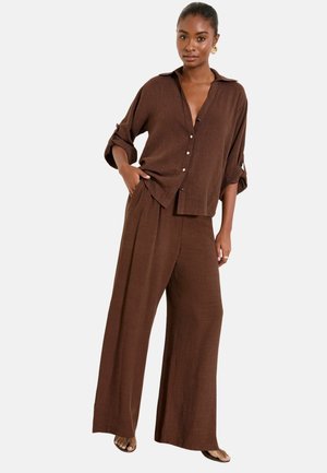 Frau steht und trägt braune, lockere, weit geschnittene Hosen und ein passendes langärmeliges Hemd mit hochgekrempelten Ärmeln.