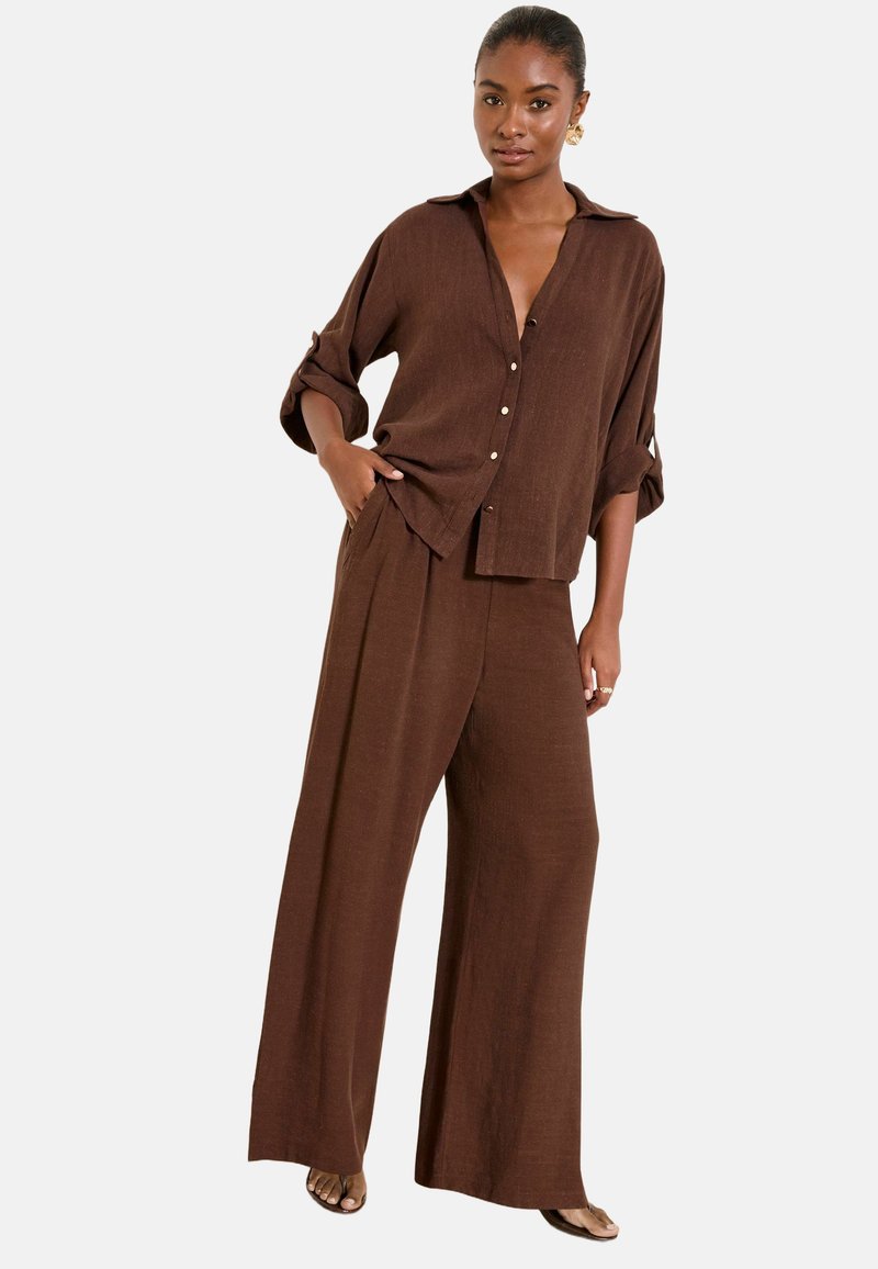 Donna in piedi che indossa pantaloni marroni larghi a gamba larga e una camicia abbottonata a maniche lunghe abbinate con le maniche arrotolate.