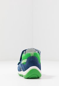 Sininen samettikenkä vihreillä verkkoaksentilla, valkoisella kumipohjalla ja säädettävällä hihnalla. Siinä on ilmanvaihtoaukkoja sivuilla.