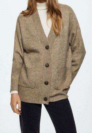 Femme portant un cardigan beige boutonné avec des poches avant par-dessus un col roulé blanc et un pantalon noir, debout avec une main dans la poche.