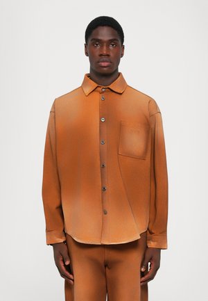 Man som bär en löst sittande, bränd orange skjorta med knappar och en bröstficka, i kombination med matchande byxor, står mot en enkel bakgrund.