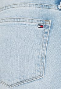 Vaaleansininen farkkutasku, jossa on näkyvät ompeleet. Taskun lähellä on pieni Tommy Hilfiger -logo punaisena, valkoisena ja sinisenä.