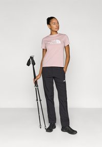 Ροζ ανοιχτόχρωμο κοντομάνικο μπλουζάκι με το λογότυπο "The North Face", συνδυασμένο με μαύρο παντελόνι εργασίας και μαύρα παπούτσια πεζοπορίας, κρατώντας μπαστούνια πεζοπορίας.