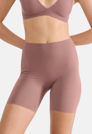 Kobieta ubrana w bezszwowe, wysokopodparte fioletowe rowerowe szorty oraz dopasowany stanik, stojąca z rękami za plecami na jednolitym tle.