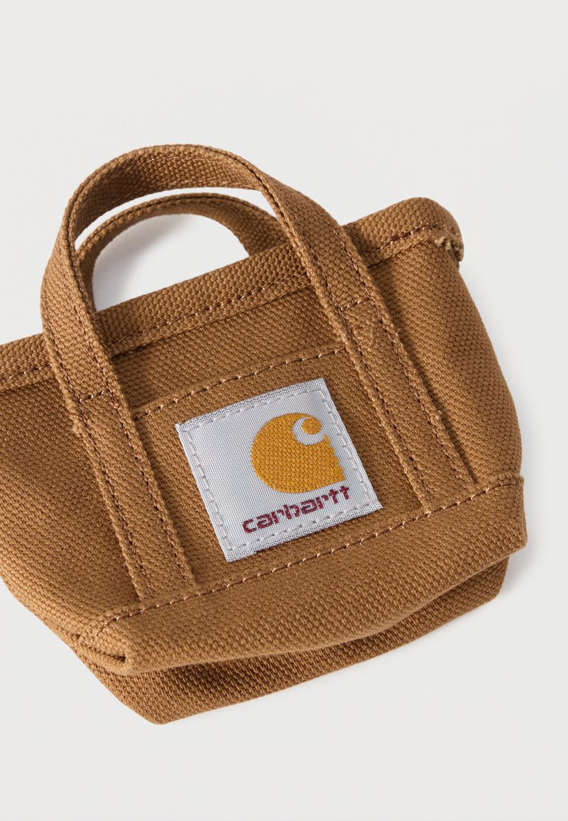 Piccola borsa a tracolla in tela marrone con doppi manici e un'etichetta bianca con un logo giallo e la scritta "carhartt".