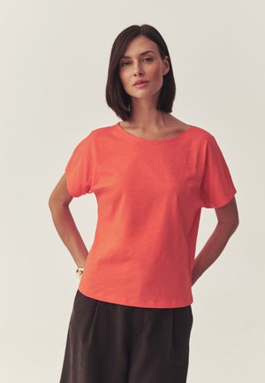 Kobieta o prostych, ciemnych włosach, ubrana w luźny, krótki pomarańczowy top i ciemne, szerokie spodnie, stoi z rękami za plecami.