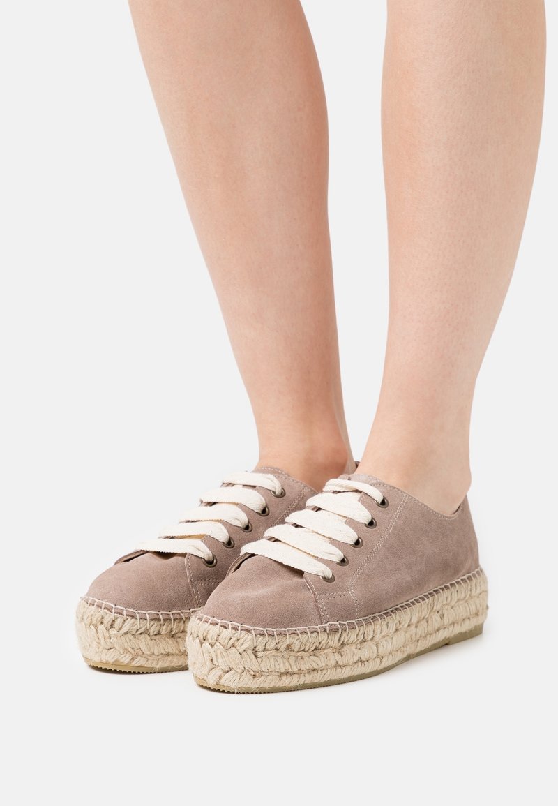 Pieds chaussés de baskets espadrilles compensées en daim taupe avec des semelles épaisses en jute tressée et des lacets blancs sur un fond uni.