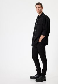 Musta ylisuuri napillinen paita rintataskuilla, yhdistettynä slim fit mustiin housuihin ja mustiin nilkkabootseihin. Liukas rakenne, minimalistinen muotoilu.